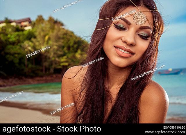 Closeup portrait of beautiful brunette woman with gold head jewelry smiling over sea tropical background