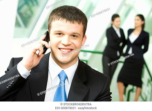 Portrait of a young successful businessman making business call on mobile looking at camera on the background of two standing women