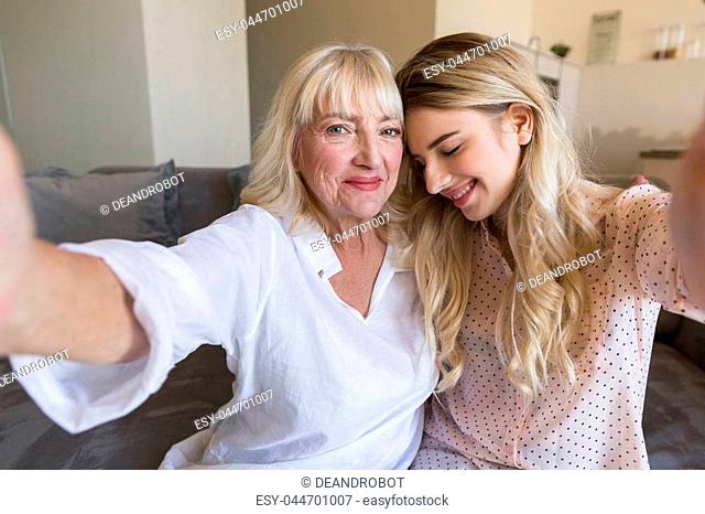 Smiling young granddaughter and her grandmother taking a selfie while sitting on a couch at home