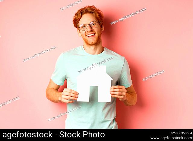 Real estate. Cheerful young man with red hair, wearing glasses and t-shirt, showing paper house cutout and smiling, buying apartment, pink background