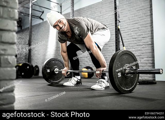 Smiling guy with a beard prepares to raise up a barbell in the gym on the gray brick wall background. He wears sportswear, white sneakers and a white cap