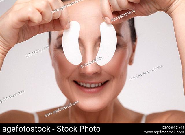 Patch Under Eyes. Beautiful middle aged woman with perfect skin showing white hydrogel patches with lifting anti-wrinkle collagen effect, gray background