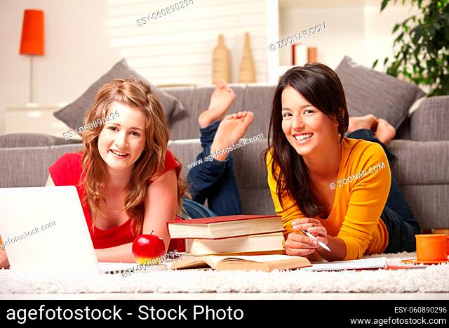 Happy teen girls lying on floor studying with laptop and books smiling at camera at home