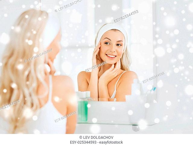 beauty, skin care and people concept - smiling young woman in hairband touching her face and looking to mirror at home bathroom over snow