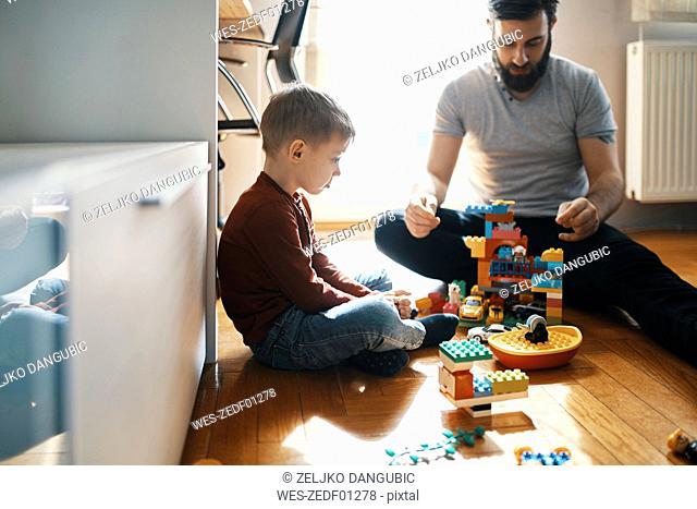 Father playing with building bricks on the floor at home ignoring his sad little son sitting beside him