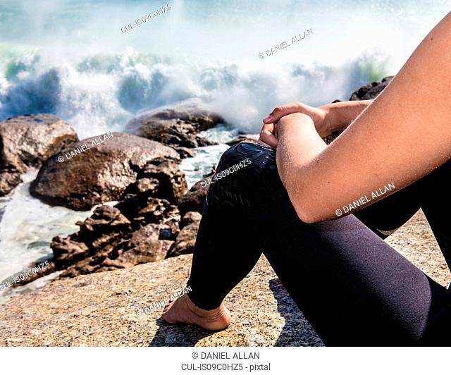 Young female surfer sitting on beach rock amongst ocean waves, cropped, Cape Town, Western Cape, South Africa