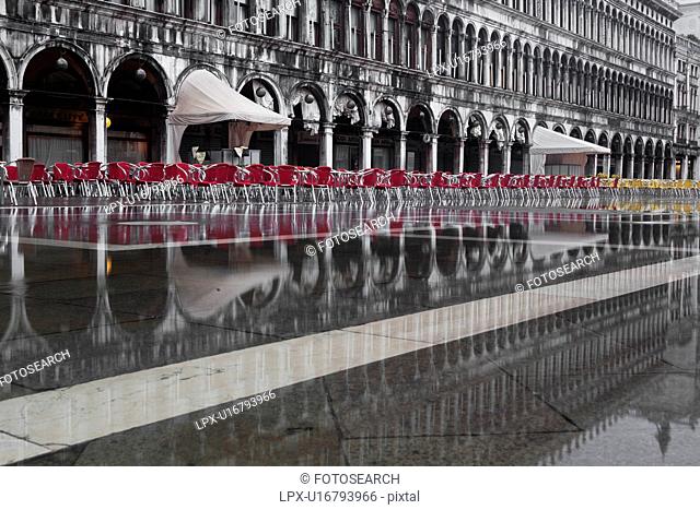 Aqua Alta flooding in Piazza San Marco: desaturated image highlighting empty red and yellow chairs, and grey facade and loggia of the Procuratie behind