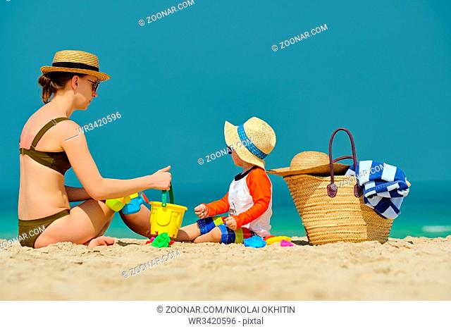 Two year old toddler boy in sun hat playing with mother on beach