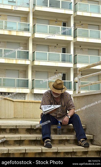 Man with hat reading newspaper on stairs in front of high-rise building, Villaricos, Andalucia, Spain, Europe