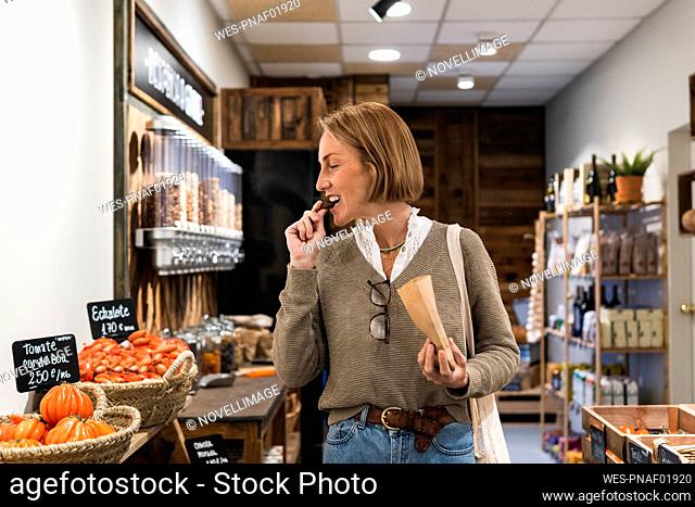 Blond woman eating food while looking at vegetables in supermarket