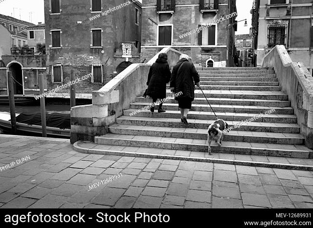 Two old women walk an old dog up the stone steps of a stone bridge in the Jewish Ghetto area Cannaregio in Venice