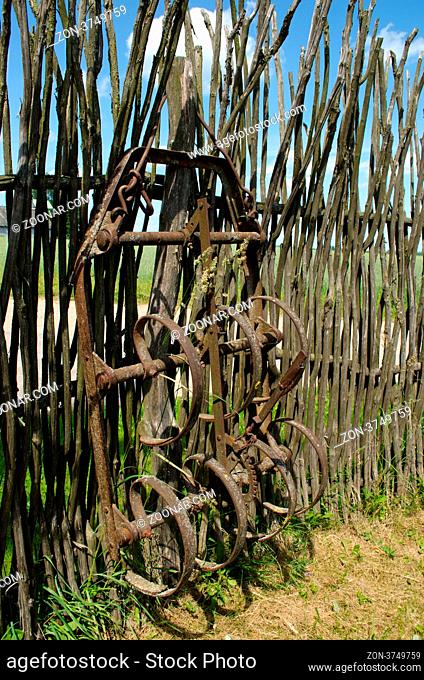 Old corroded harrowing field the tool stand near handmade fence from tree branches