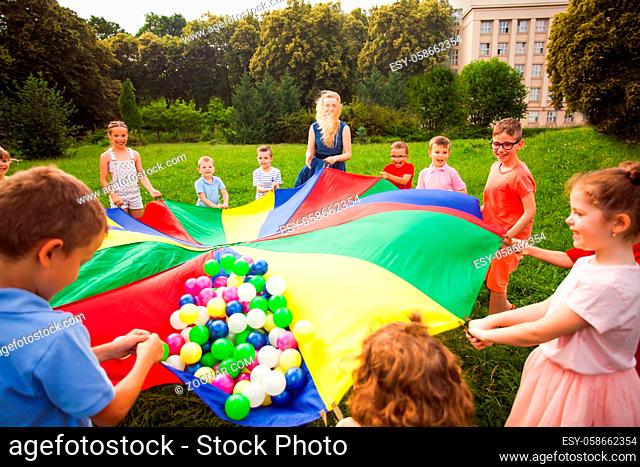 Kids with their educator waving large rainbow cloth outdoors in the city park. Outdoor social anti-bullying lesson. Teaching to cooperate in summer daycamp