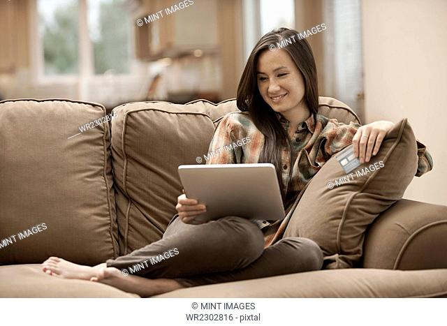 A woman sitting on a sofa at home holding a digital tablet and a credit card, shopping online