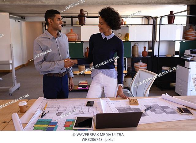 Front view of happy mixed-race business people shaking hands with plans in foreground on desk