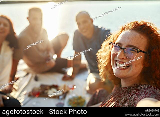 Happy redheaded woman having picnic with friends on jetty at a lake at sunset