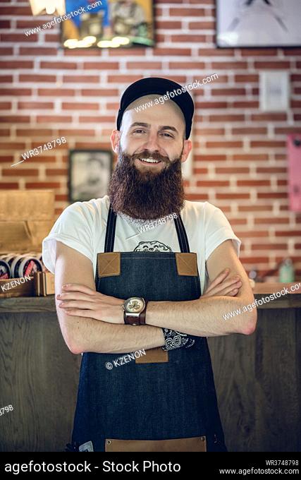 Portrait of a cool bearded hairstylist wearing wristwatch, apron and cap while looking at camera with confidence and passion for his job