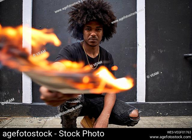 Portrait of young man crouching on pavement holding burning newspaper