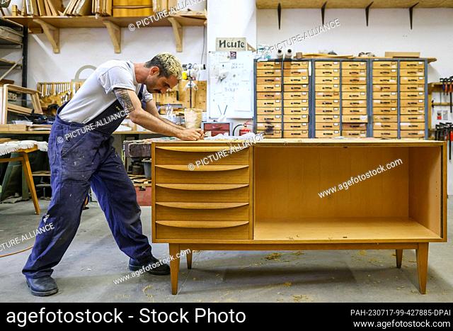 dpatop - PRODUCTION - 10 July 2023, Saxony, Leipzig: Kris Sullivan removes old varnish from a sideboard ""Model 602k"" from Deutsche Werkstätten Hellerau