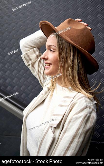 Smiling woman holding hat while looking away against wall