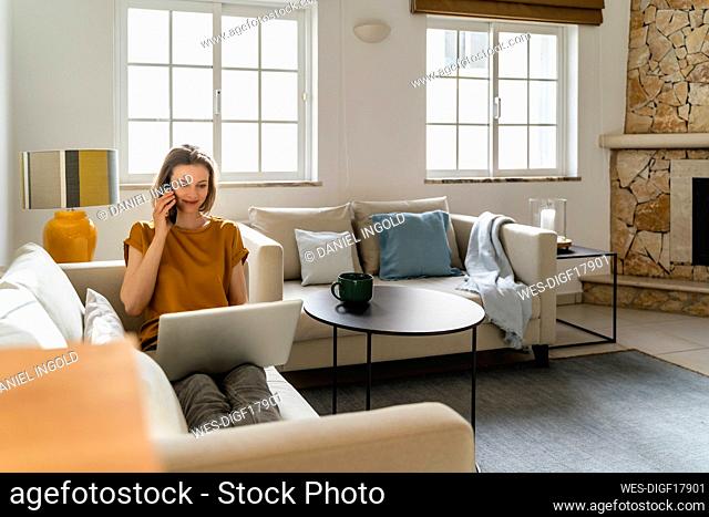 Smiling freelancer talking on smart phone sitting with laptop working at home