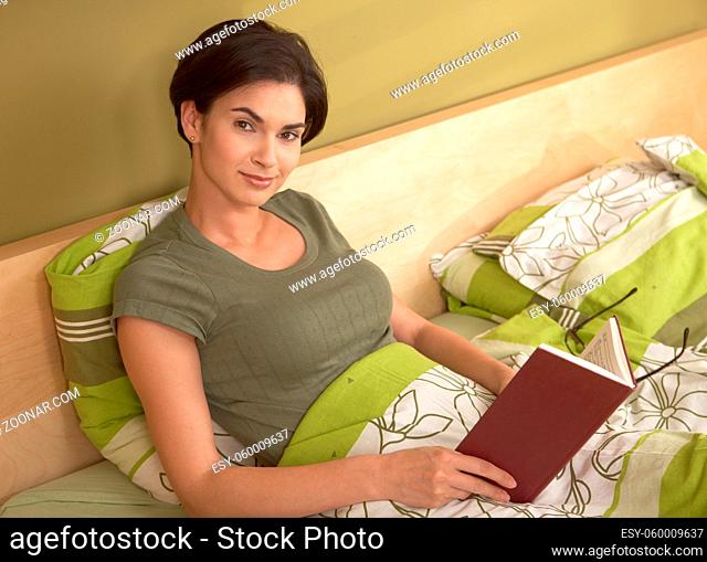 Portrait of smiling woman reading in bed, looking at camera, holding book
