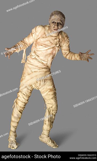 Man dressed up as a mummy for Halloween making stiff arm movements against a gray background