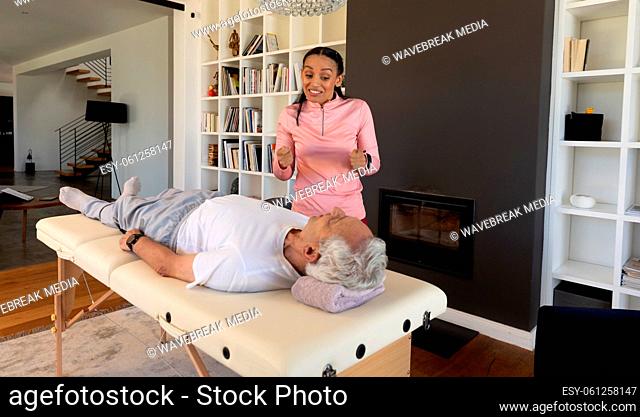 Biracial female physiotherapist gesturing while guiding caucasian senior man lying on table at home
