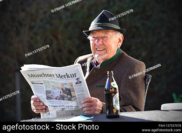 Ruestiger old man (92 years) reads a daily newspaper (Muenchner Merkur) on November 22nd, 2020. He wears traditional Bavarian clothes - next to him is a bottle...
