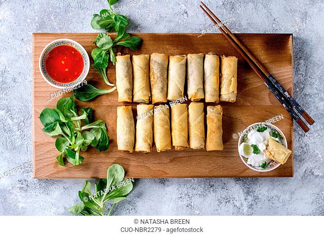 Fried spring rolls with red and white sauces, served on wood serving board with fresh green salad and wooden chopsticks over gray blue texture background