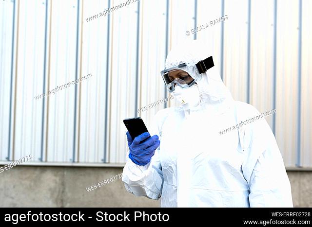 Female scientist wearing protective suit and mask and looking at smartphone