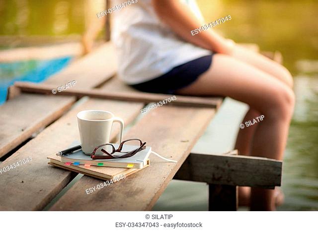 Notebooks, pen, glasses, and coffee cup are putting down on the wood waterside while young woman sitting beside them in morning time on weekend