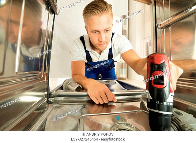 Young Man In Overall Repairing Dishwasher With Electric Drill