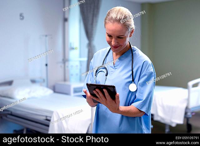 Smiling caucasian female doctor in hospital wearing scrubs and stethoscope using tablet