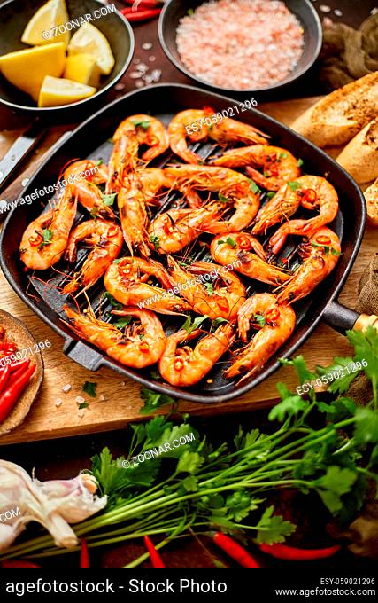 Roasted tiger prawns on iron grilling pan with fresh parsley, lemon, chilli and bread over rusty background, top view, flat lay