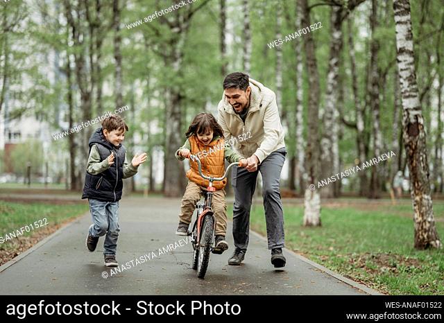 Boy cheering brother learning to ride bicycle with father at park