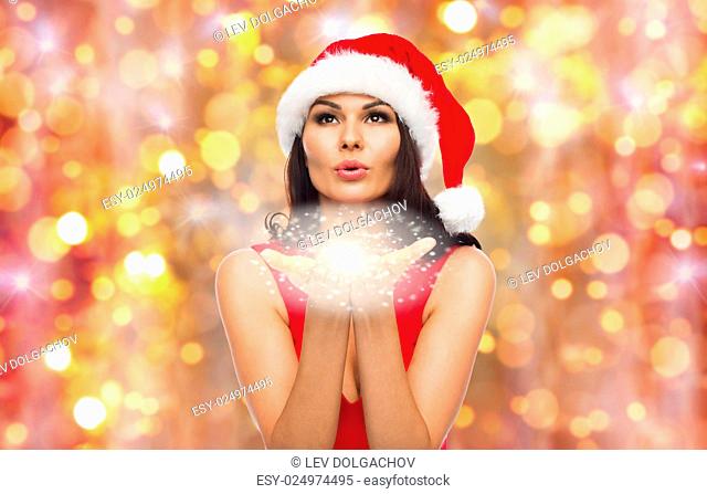 people, holidays, christmas and celebration concept - beautiful sexy woman in santa hat and red dress blowing fairy dust or snowflakes off her palms over lights...
