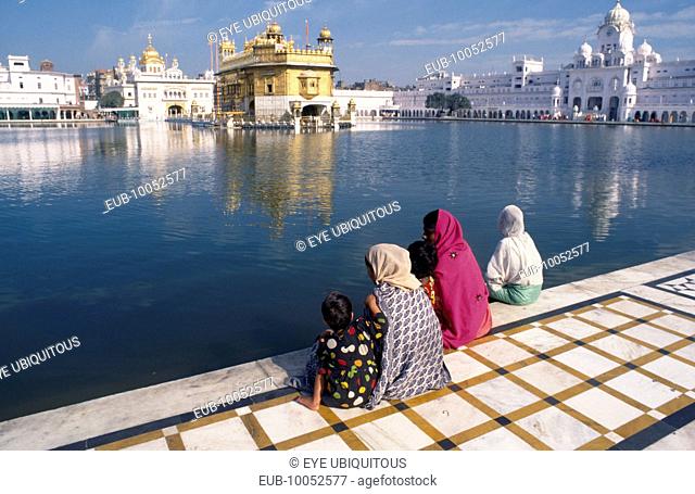 Three women and children sitting on marble walkway beside the sacred pool looking towards The Golden Temple