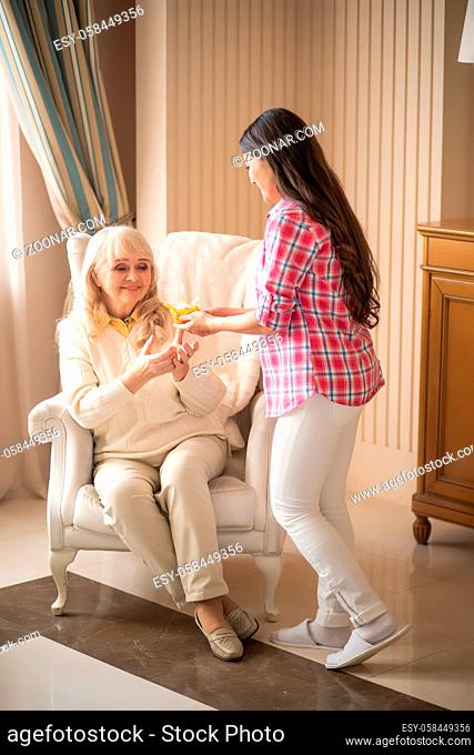 Vertical Shot Of Caring Daughter Congratulating Her Senior Mother With A Birthday Cupcake. Older Woman Is Sitting On A While Chair And Takes Birthday Cake