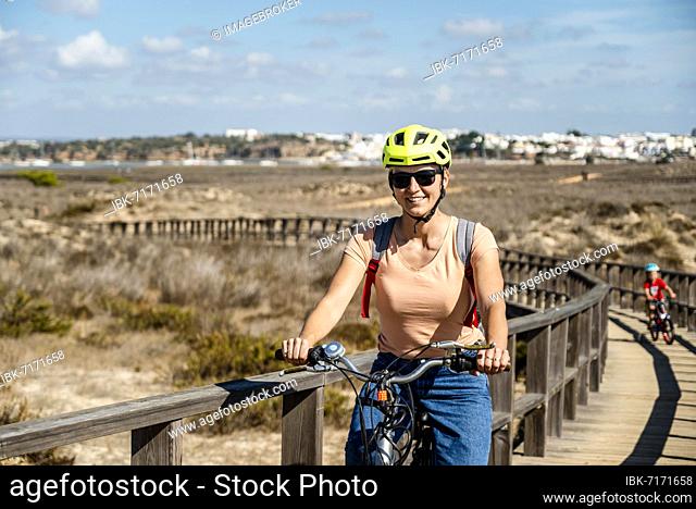 Mother riding a bicycle with her son on wooden walkways in Alvor, Algarve, Portugal, Europe