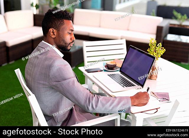 African american man sitting in a cafe using laptop and writing in notebook