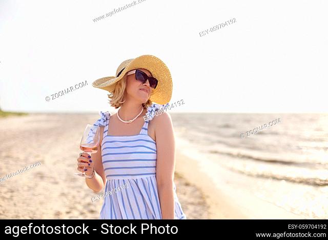 Glad adult female in summer outfit smiling and looking away while enjoying wine on sandy beach near sea