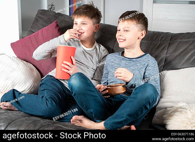 Smiling school aged boys sit on sofa, eat popcorn and watch comic TV show. Happy children relaxing at home. Kids having fun in domestic room at daytime together