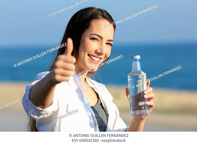 Portrait of a happy girl holding a water bottle gesturing thumbs up on the beach