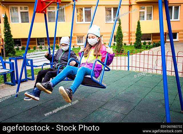 The elementary school boy and girl are wearing a medical masks and swing on a swing. The serious children rest in the playground in the fall