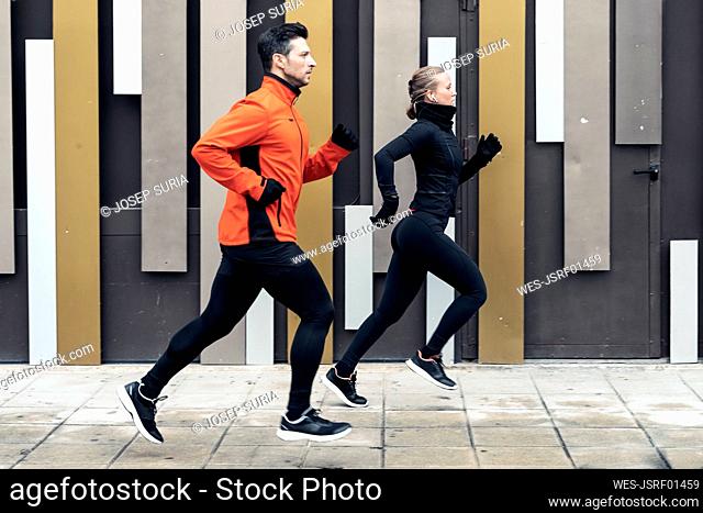 Determined male and female friends jogging on footpath by wall