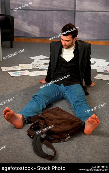 Trendy office worker sitting on office floor bare feet, surrounded with papers, documents and laptop bag