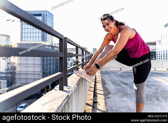 Smiling young woman tying shoelace on railing against sky