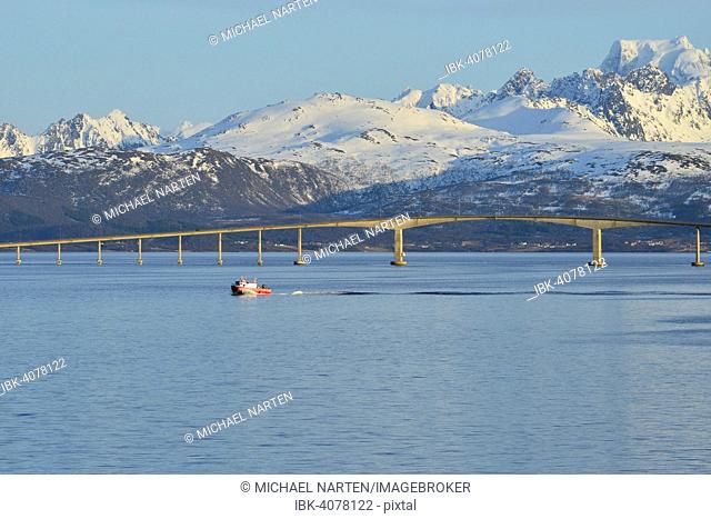 Red boat in front of Hadsel Bridge across the strait, at the back the snow-capped mountains of Hinnøya island, Langøysund, near Stokmarknes, Nordland