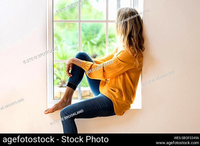 Woman sitting on windowsill while looking through window in living room
